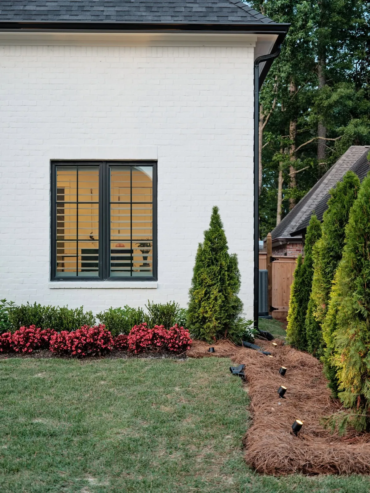 White brick home with landscape lights, red flowering beds and tall evergreens
