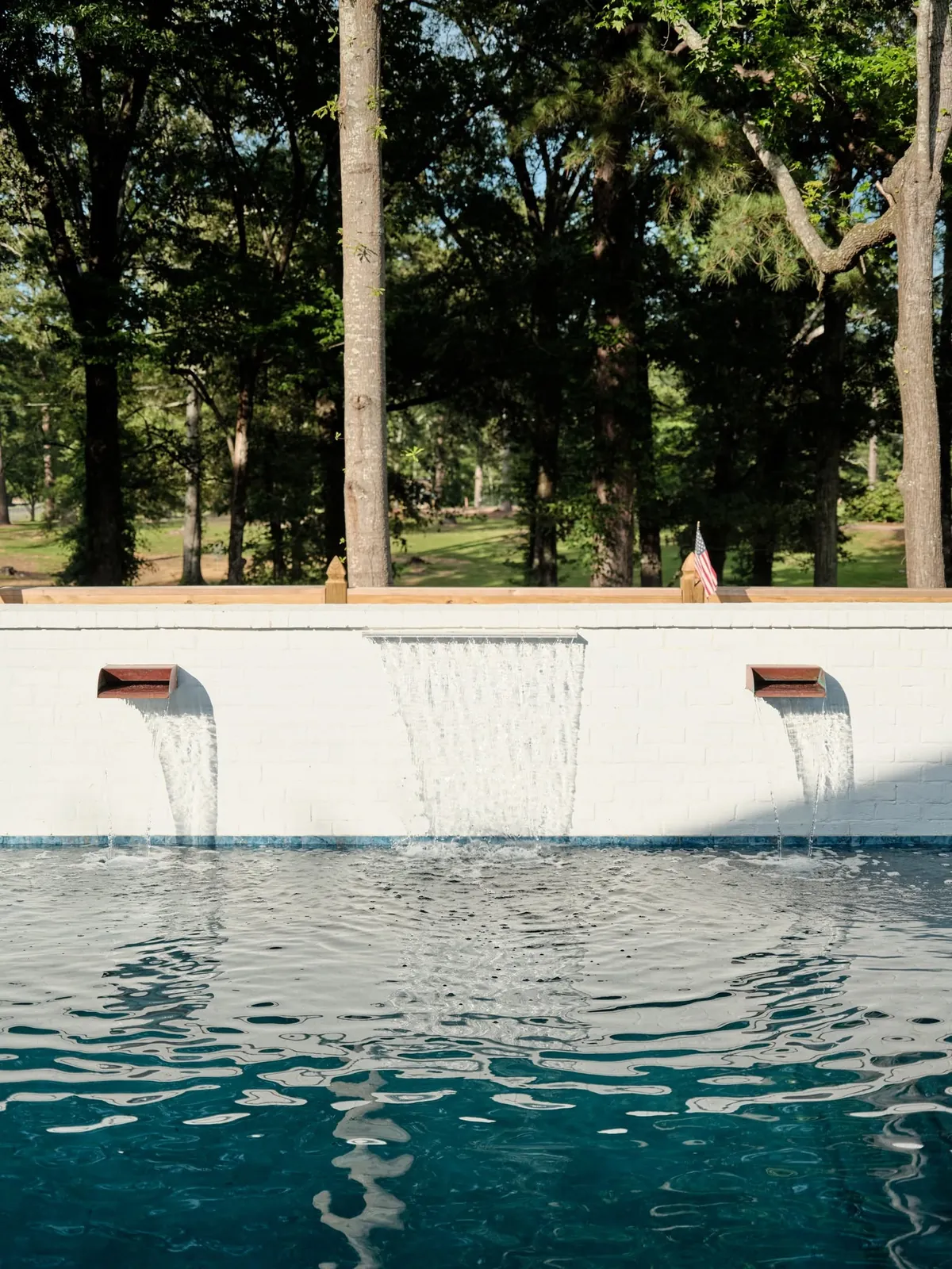 Pool with a white wall and spillways pouring water into it under tall trees